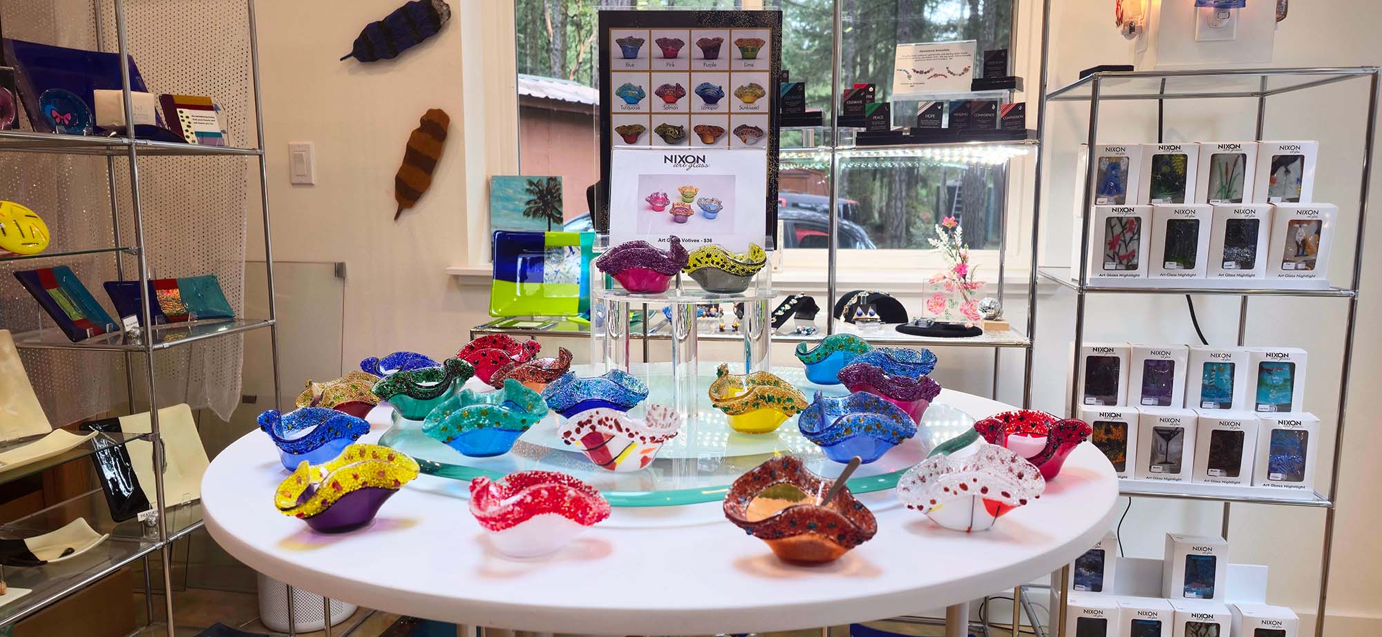 Table displaying colorful glass art pieces in a store setting with shelves and displays in the background.